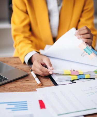 Mulher organizando papéis e documentos em sua mesa, com notebook e calculadora, revisando informações financeiras.