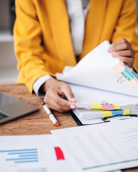 Mulher organizando papéis e documentos em sua mesa, com notebook e calculadora, revisando informações financeiras.