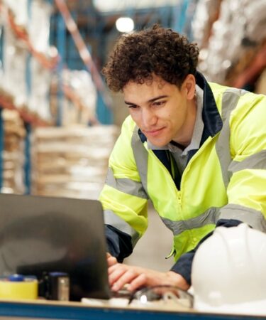 homem agachado mexe em um laptop em um deposito.