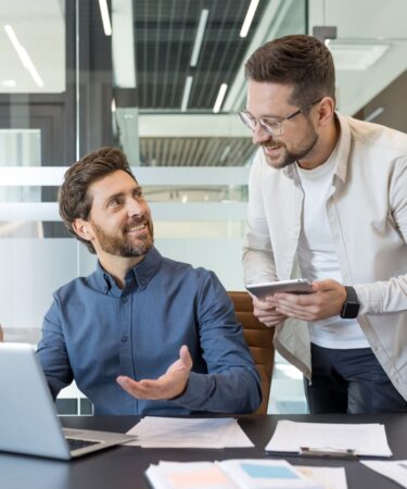 Dois jovens profissionais, em um ambiente moderno e corporativo, conversam em frente a um computador enquanto sorriem amistosamente.