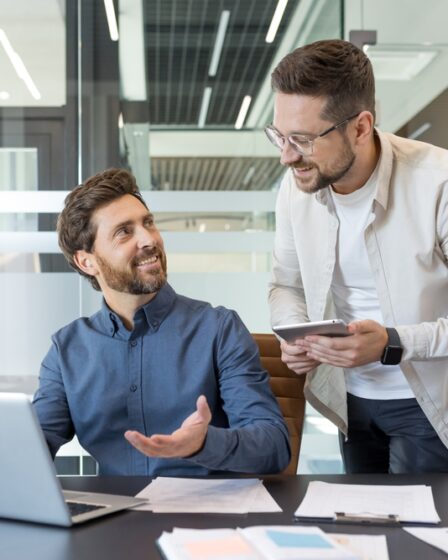 Dois jovens profissionais, em um ambiente moderno e corporativo, conversam em frente a um computador enquanto sorriem amistosamente.