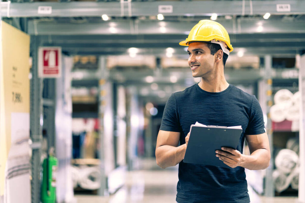 Técnico em Logística com capacete de segurança e prancheta realizando conferência de estoque em um centro de distribuição. 