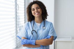 Técnica de enfermagem sorrindo com estetoscópio e luvas em ambiente clínico, demonstrando preparo para o atendimento aos pacientes.