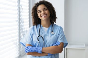 Técnica de enfermagem sorrindo com estetoscópio e luvas em ambiente clínico, demonstrando preparo para o atendimento aos pacientes.
