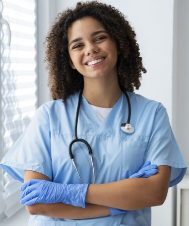 Técnica de enfermagem sorrindo com estetoscópio e luvas em ambiente clínico, demonstrando preparo para o atendimento aos pacientes.
