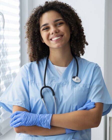Técnica de enfermagem sorrindo com estetoscópio e luvas em ambiente clínico, demonstrando preparo para o atendimento aos pacientes.