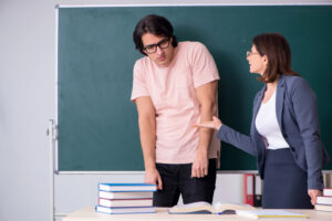 Professora repreende aluno em sala de aula. Há uma mesa no primeiro plano com cadernos em cima. No fundo, um quadro negro de cor verde.
