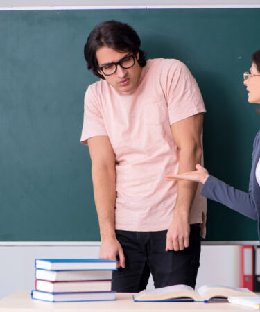 Professora repreende aluno em sala de aula. Há uma mesa no primeiro plano com cadernos em cima. No fundo, um quadro negro de cor verde.