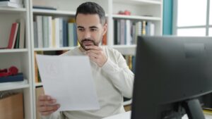 Homem branco em frente ao computador em ambiente de biblioteca, analisando documento.