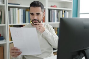 Homem branco em frente ao computador em ambiente de biblioteca, analisando documento.