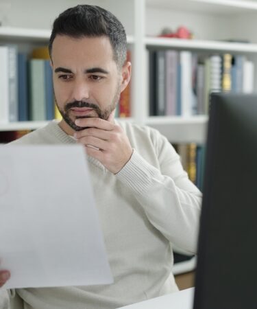 Homem branco em frente ao computador em ambiente de biblioteca, analisando documento.