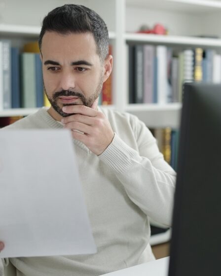 Homem branco em frente ao computador em ambiente de biblioteca, analisando documento.