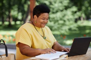 Mulher negra idosa usando laptop ao ar livre em uma mesa de café e sorrindo.