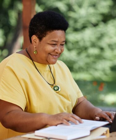 Mulher negra idosa usando laptop ao ar livre em uma mesa de café e sorrindo.