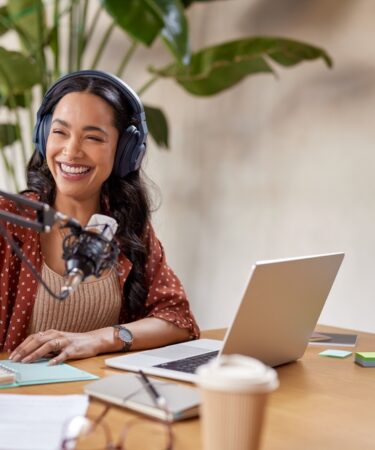 Mulher jovem usando fone de ouvido sorri e faz anotações em uma agenda. Ela está sentada com um notebook na mesa em sua frente onde há outros objetos como um copo, um óculos e papeis. Além disso, há um microfone preso em um pedestal próximo a sua boca.