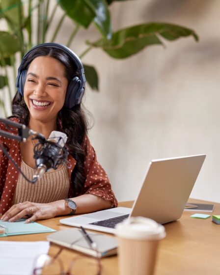 Mulher jovem usando fone de ouvido sorri e faz anotações em uma agenda. Ela está sentada com um notebook na mesa em sua frente onde há outros objetos como um copo, um óculos e papeis. Além disso, há um microfone preso em um pedestal próximo a sua boca.
