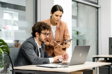 Homem de óculos e terno está sentado mexendo em seu laptop enquanto uma mulher vestindo blusa bege e segurando uma prancheta conversa com ele.