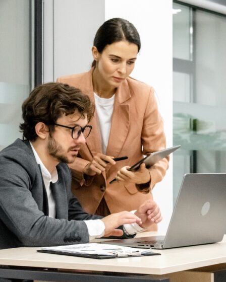 Homem de óculos e terno está sentado mexendo em seu laptop enquanto uma mulher vestindo blusa bege e segurando uma prancheta conversa com ele.