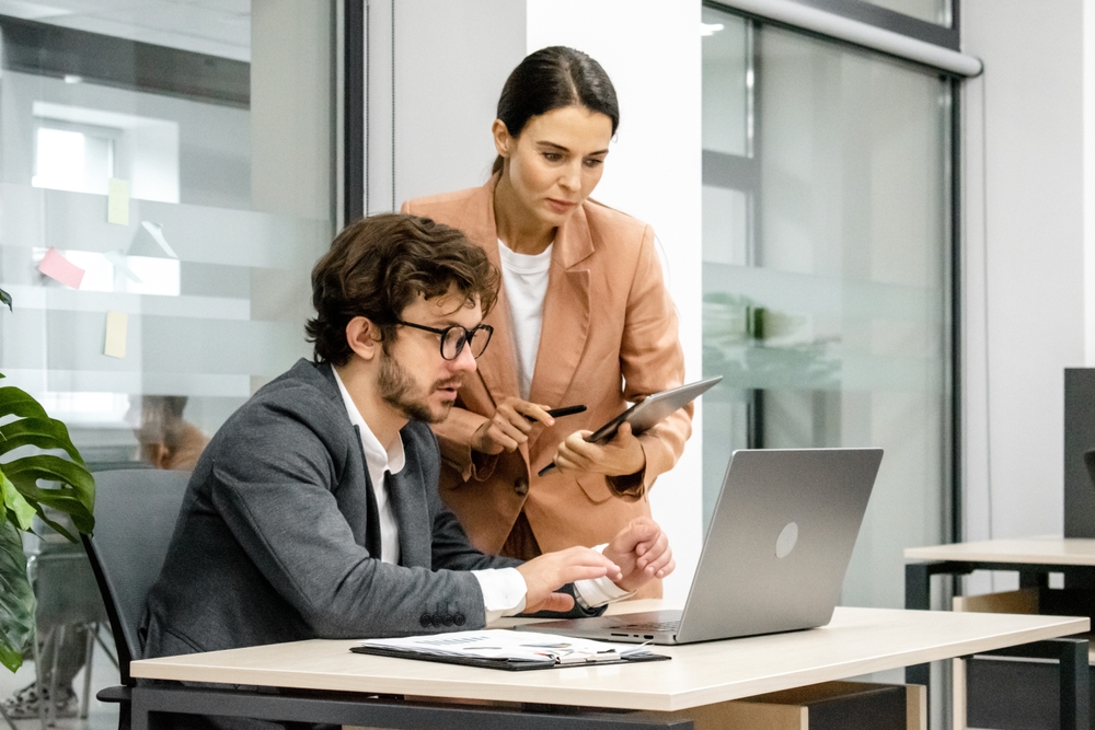 Homem de óculos e terno está sentado mexendo em seu laptop enquanto uma mulher vestindo blusa bege e segurando uma prancheta conversa com ele.
