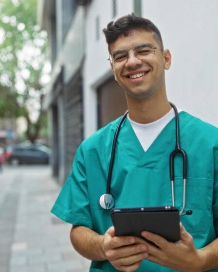 Jovem técnico de enfermagem sorrindo vestindo uniforme e estetoscópio.