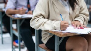 menina com blusa e saia, está sentada realizando um exame com uma caneta azul. Ao fundo, mais pessoas realizam o exame.