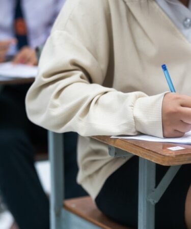 menina com blusa e saia, está sentada realizando um exame com uma caneta azul. Ao fundo, mais pessoas realizam o exame.