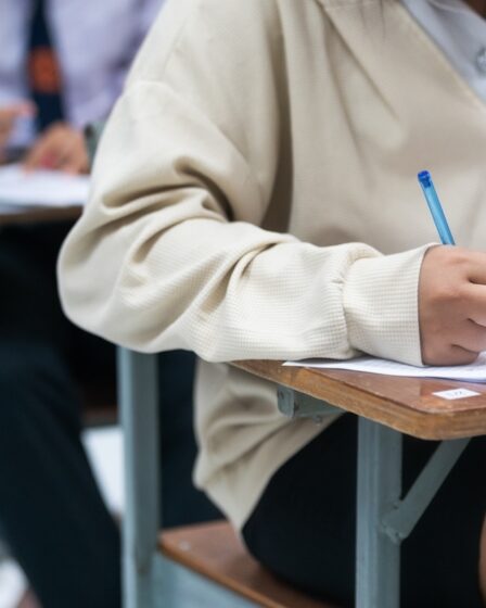 menina com blusa e saia, está sentada realizando um exame com uma caneta azul. Ao fundo, mais pessoas realizam o exame.
