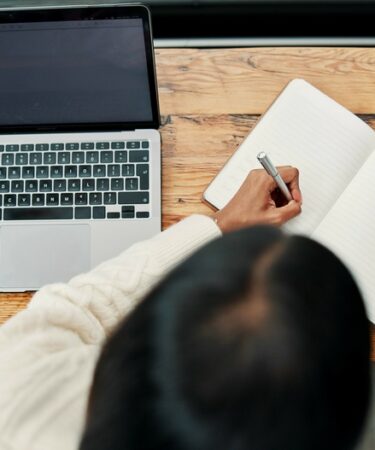 Pessoa escrevendo em caderno sobre mesa com café e notebook.