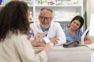 Estudante do curso de farmácia sendo atendida no balcão por farmacêutico experiente, enquanto segura uma cartela de comprimidos.