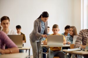 Mulher jovem de pé ao lado de um menino adolescente o orienta apontando para a tela de um laptop. Ao redor, há diversos estudantes adolescentes sentados em suas carteiras.