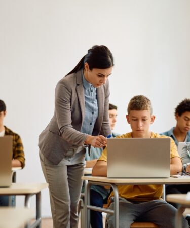 Mulher jovem de pé ao lado de um menino adolescente o orienta apontando para a tela de um laptop. Ao redor, há diversos estudantes adolescentes sentados em suas carteiras.
