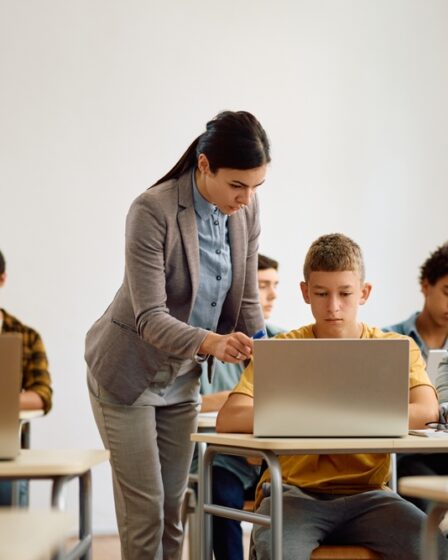 Mulher jovem de pé ao lado de um menino adolescente o orienta apontando para a tela de um laptop. Ao redor, há diversos estudantes adolescentes sentados em suas carteiras.