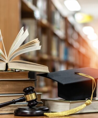 Há duas colunas de livros empilhados. Na primeira, cinco livros estão empilhados e o último está aberto. Na segunda, há apenas um livro e em cima dele um capelo. Na frente dos livros há um malhete. O fundo é desfocado com raios de luz solar ao fundo.