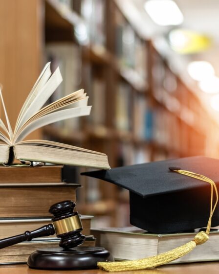 Há duas colunas de livros empilhados. Na primeira, cinco livros estão empilhados e o último está aberto. Na segunda, há apenas um livro e em cima dele um capelo. Na frente dos livros há um malhete. O fundo é desfocado com raios de luz solar ao fundo.