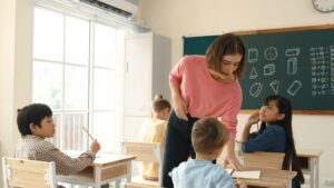 Professora caucasiana caminhando e verificando a lição de casa dos alunos na sala de aula, enquanto crianças de diferentes origens étnicas realizam atividades escolares. Alunos inteligentes de diferentes origens étnicas assistindo à aula e fazendo anotações em uma escola primária. Pedagogia.