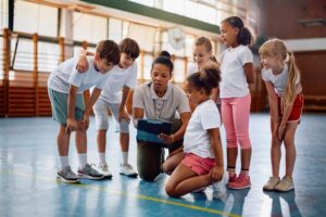Professora de educação física orienta grupo de crianças durante atividade esportiva em quadra esportiva escolar.