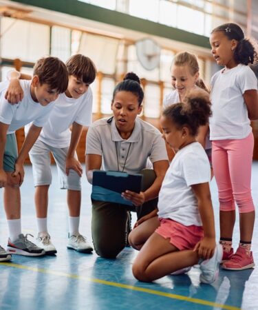 Professora de educação física orienta grupo de crianças durante atividade esportiva em quadra esportiva escolar.
