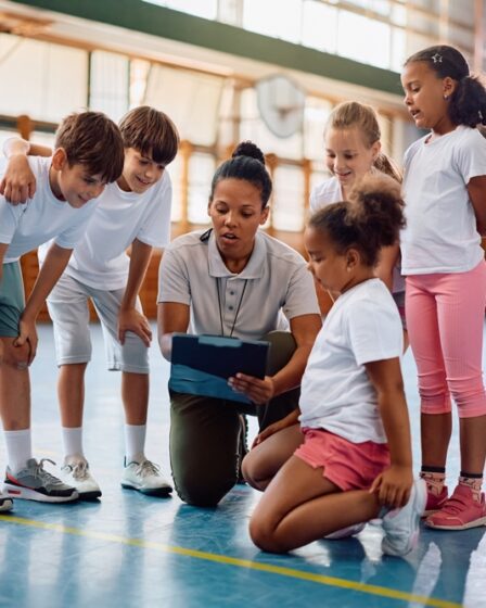 Professora de educação física orienta grupo de crianças durante atividade esportiva em quadra esportiva escolar.