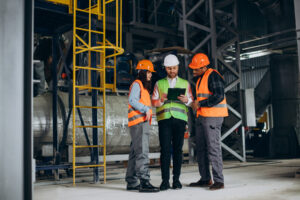Profissionais em ambiente industrial usando capacetes e coletes, representando práticas de Segurança do Trabalho no local.