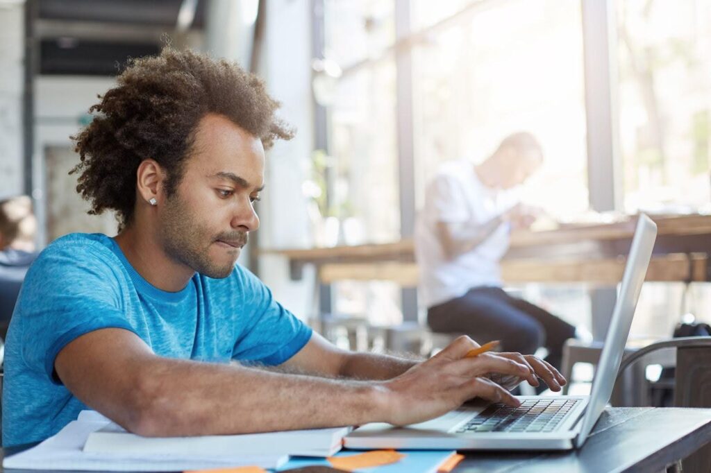 Homem jovem focado em seu notebook em um ambiente de cafeteria, consulta livros, buscando entender o que é ABNT para organizar suas referências bibliográficas.