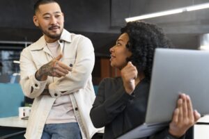 Colegas de trabalho discutem projeto ao lado de um notebook, utilizando comunicação assertiva para alinhar tarefas e decisões.
