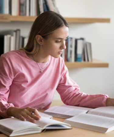 Mulher estudante concentrada lendo e fazendo anotações em ambiente de estudo.