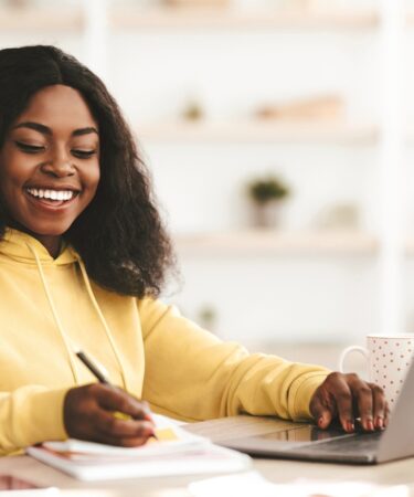 Mulher jovem negra, vestindo um moletom amarelo claro, está estudando em uma mesa com livros e um notebook.