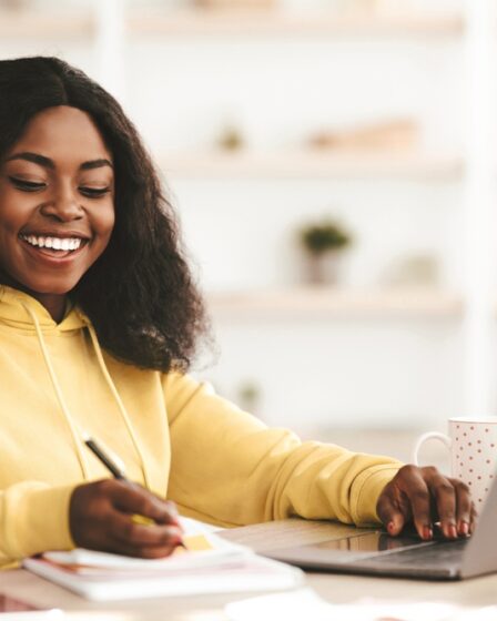 Mulher jovem negra, vestindo um moletom amarelo claro, está estudando em uma mesa com livros e um notebook.