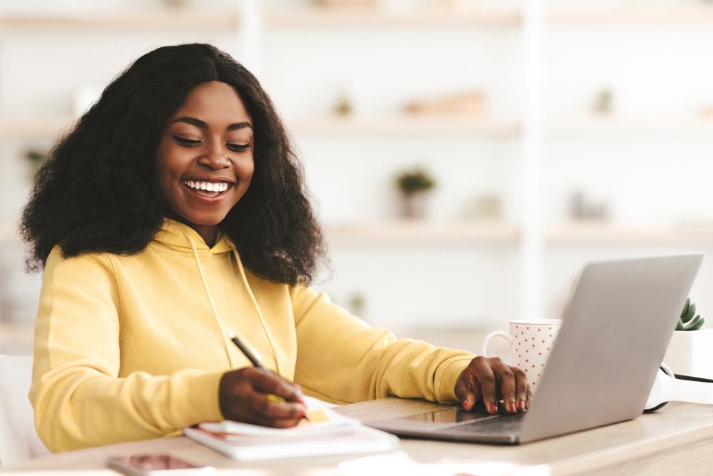 Mulher jovem negra, vestindo um moletom amarelo claro, está estudando em uma mesa com livros e um notebook.