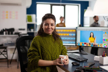 Mulher trabalhando em um escritório, segurando uma caneca enquanto edita imagens em softwares de design, cenário ideal para quem busca um curso de Design Gráfico.