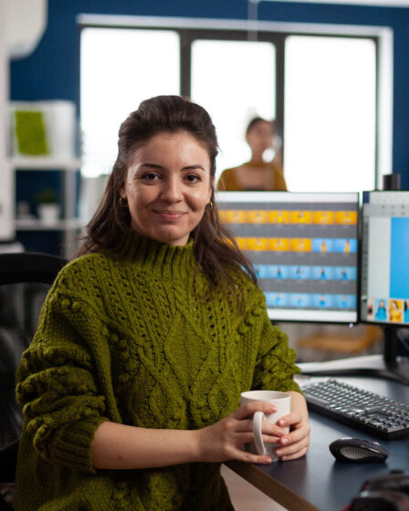 Mulher trabalhando em um escritório, segurando uma caneca enquanto edita imagens em softwares de design, cenário ideal para quem busca um curso de Design Gráfico.