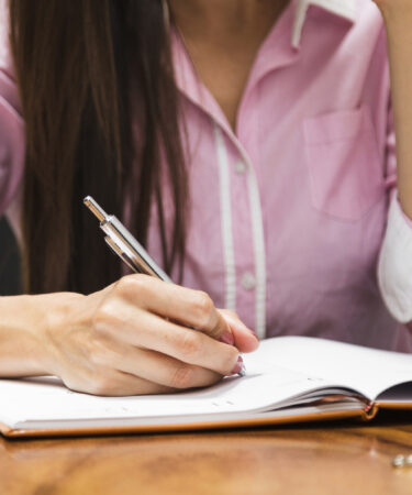 Mulher anotando ideias em um caderno sobre a mesa, simbolizando como fazer um artigo de opinião de forma organizada.