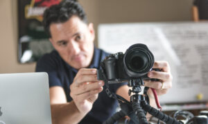 Homem jovem, profissional do audiovisual, realizando a captação de imagens com câmeras e microfone em local a céu aberto.