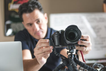 Homem jovem, profissional do audiovisual, realizando a captação de imagens com câmeras e microfone em local a céu aberto.